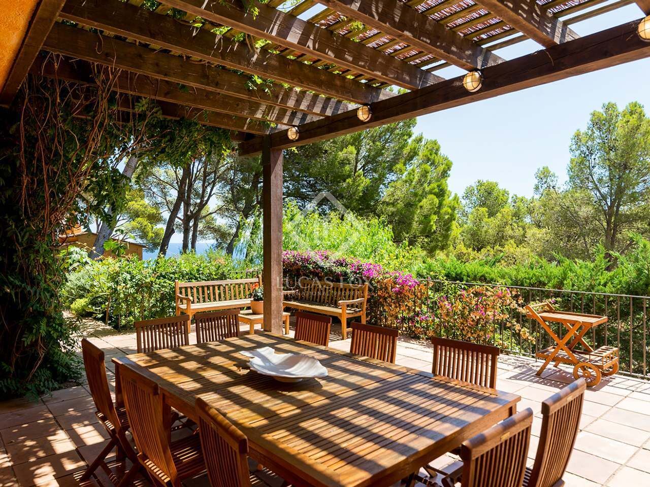 An outdoor patio with wooden furniture, including a table and benches, surrounded by lush greenery and flowering plants.