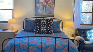Ein Bett mit einem gemusterten Überwurf, ein Blumenbild an der Wand und ein Sessel mit einer Lampe daneben.