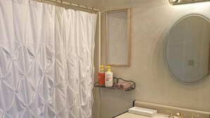 Ein Badezimmer mit weißer Duschvorhang, einem Spiegel, einem Waschbecken mit zwei Wasserhähnen und einem Ablagebereich für Toilettenartikel.