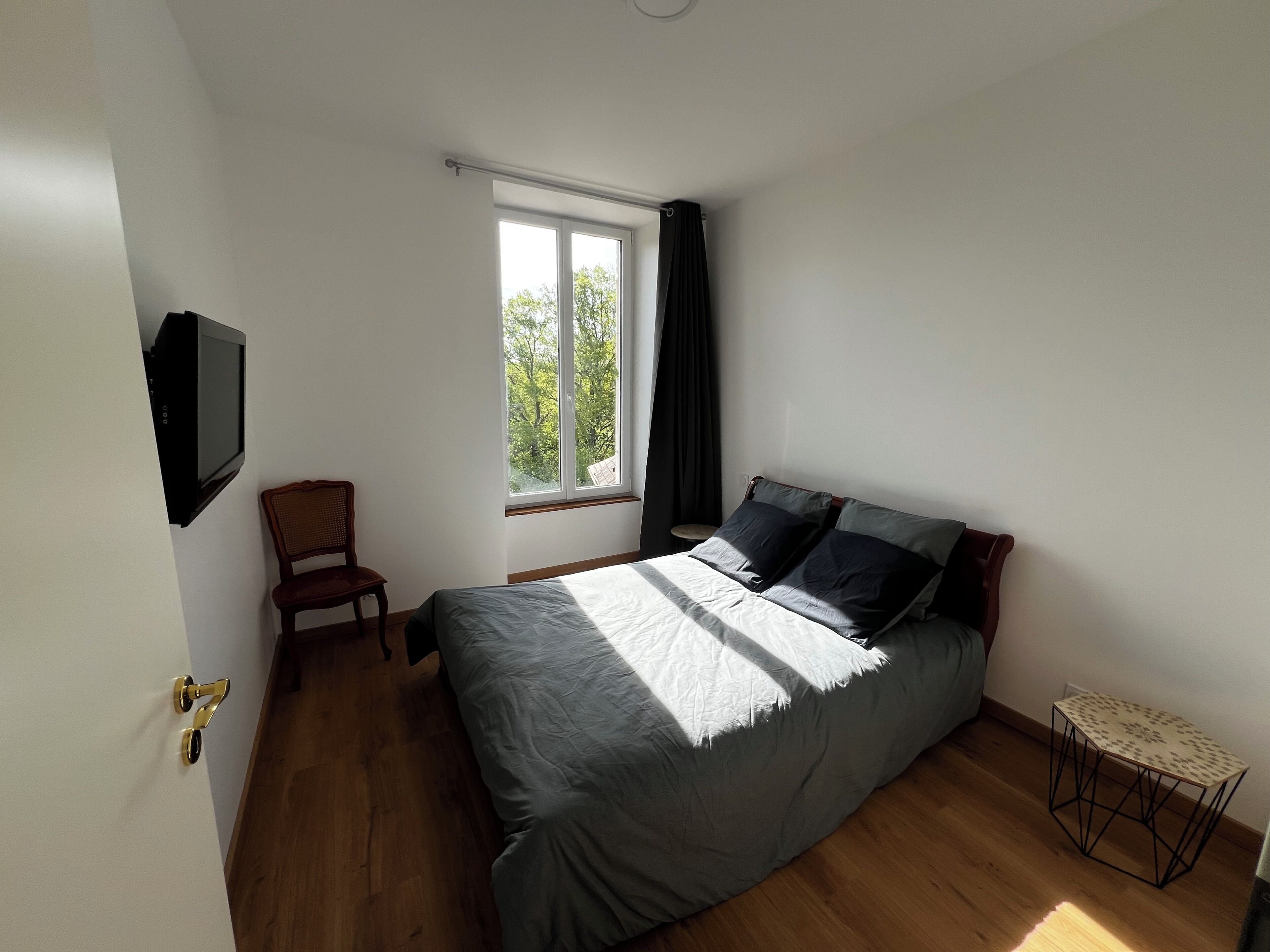 A bedroom with a bed, a chair, a small table, a TV, and a window with curtains.