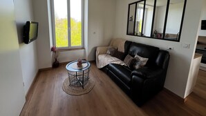 A modern living room with a black leather sofa, a small round table with a woven base, a flat-screen TV mounted on the wall, and a large window providing a view of greenery outside.