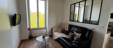 A modern living room with a black leather sofa, a small round table with a woven base, a flat-screen TV mounted on the wall, and a large window providing a view of greenery outside.