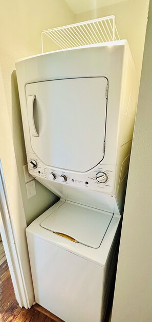 A compact, white, stacked washer and dryer unit in a corner with a metal shelf on top. - Chic Downtown Home (San Antonio)