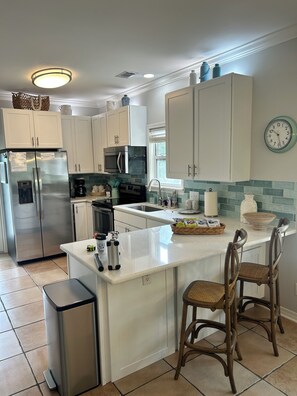 A modern kitchen with white cabinetry, a stainless steel refrigerator, a microwave, and a sink. There are two wooden chairs at the island counter. - Charming 30A Beach Home, 5Min walk to Gulf, on front of the community pool, A/C (Santa Rosa Beach)
