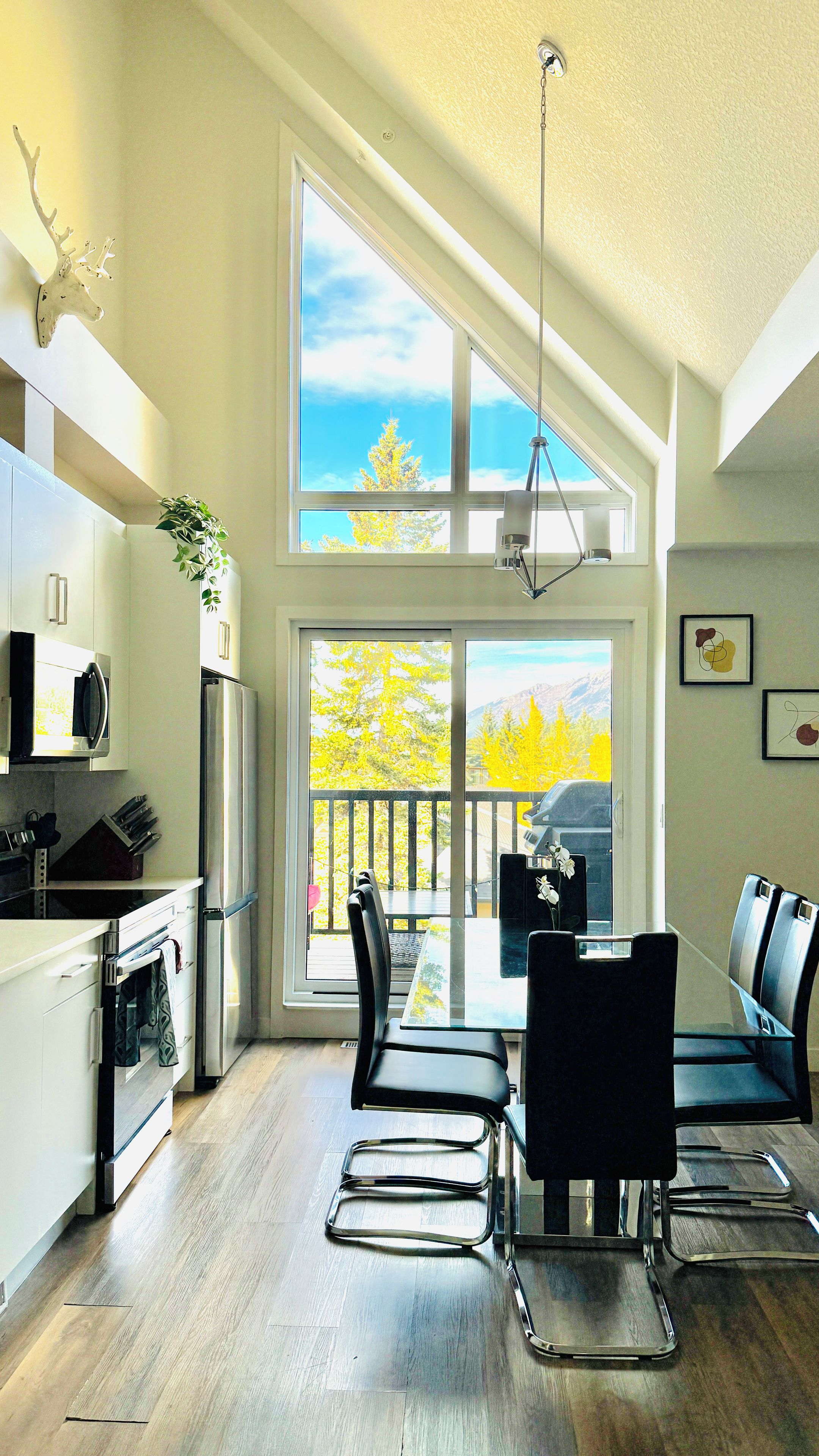 A modern kitchen with a large window, a dining area with a glass table and chairs, and a view of trees and mountains.
