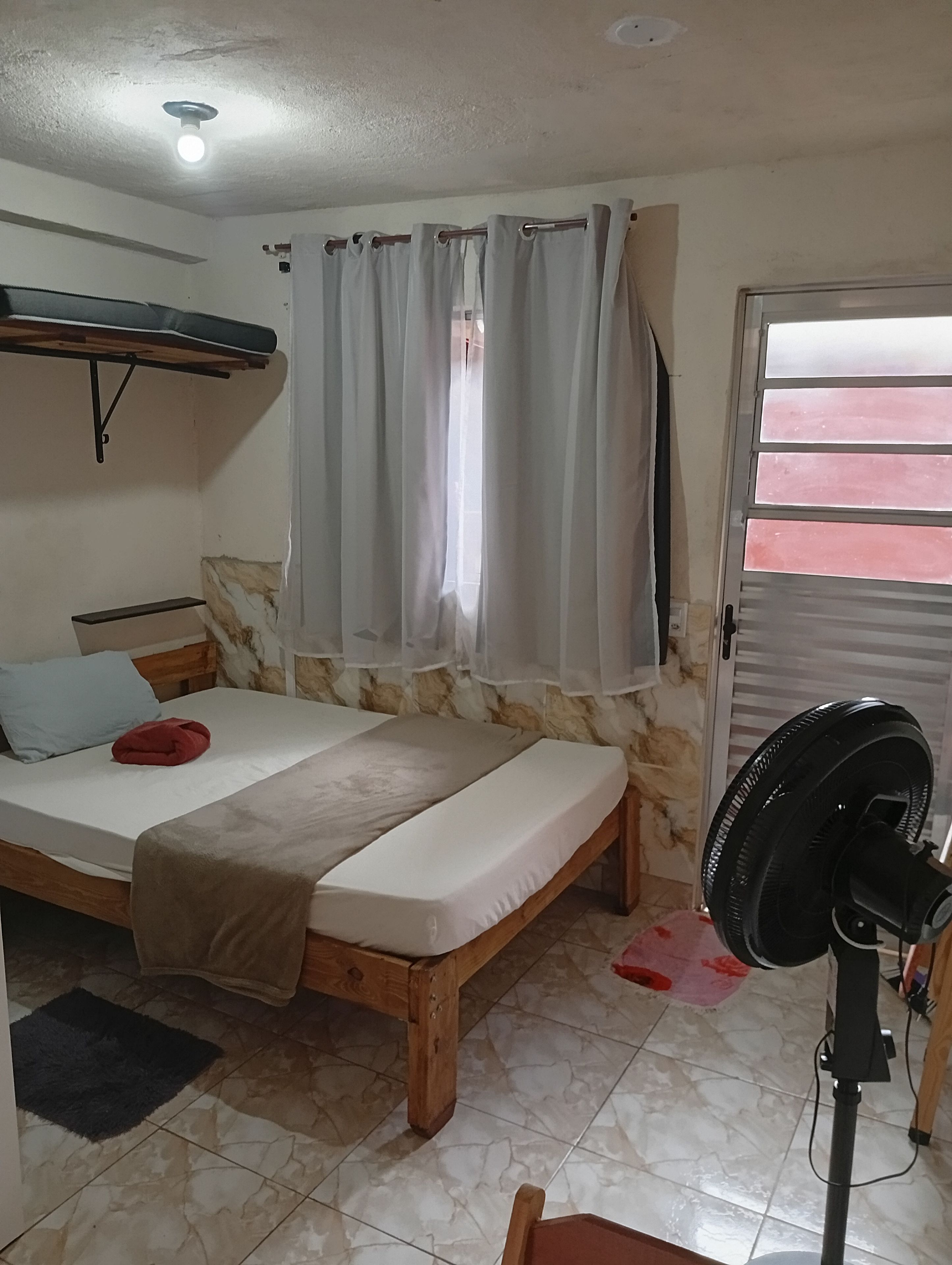 Ein Zimmer mit einem Bett, einem Ventilator, einem Fenster mit Vorhängen, einer Tür mit einem Streifenrollo und einem Regal.