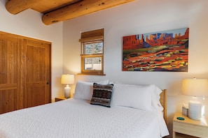 Un dormitorio con una cama grande, vigas de madera en el techo, una ventana con persianas, un cuadro en la pared y una mesita de noche con una lámpara.