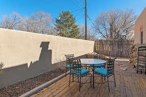 Un patio con una mesa redonda y cuatro sillas, una terraza de madera, un muro bajo y una valla de madera al fondo.