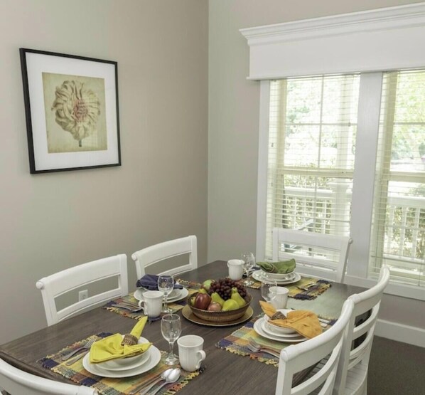 Une table à manger est dressée avec des assiettes, des verres et un saladier rempli de fruits, entourée de chaises blanches dans une pièce dotée d’une fenêtre et d’un tableau encadré au mur.