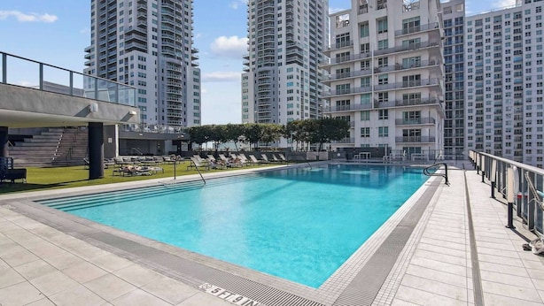 Una piscina con sillas de descanso, un paseo techado y edificios altos al fondo.