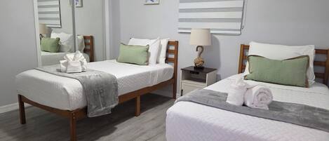 Habitación de hotel con dos camas, un espejo, una ventana con persianas y una mesita de noche con lámpara.