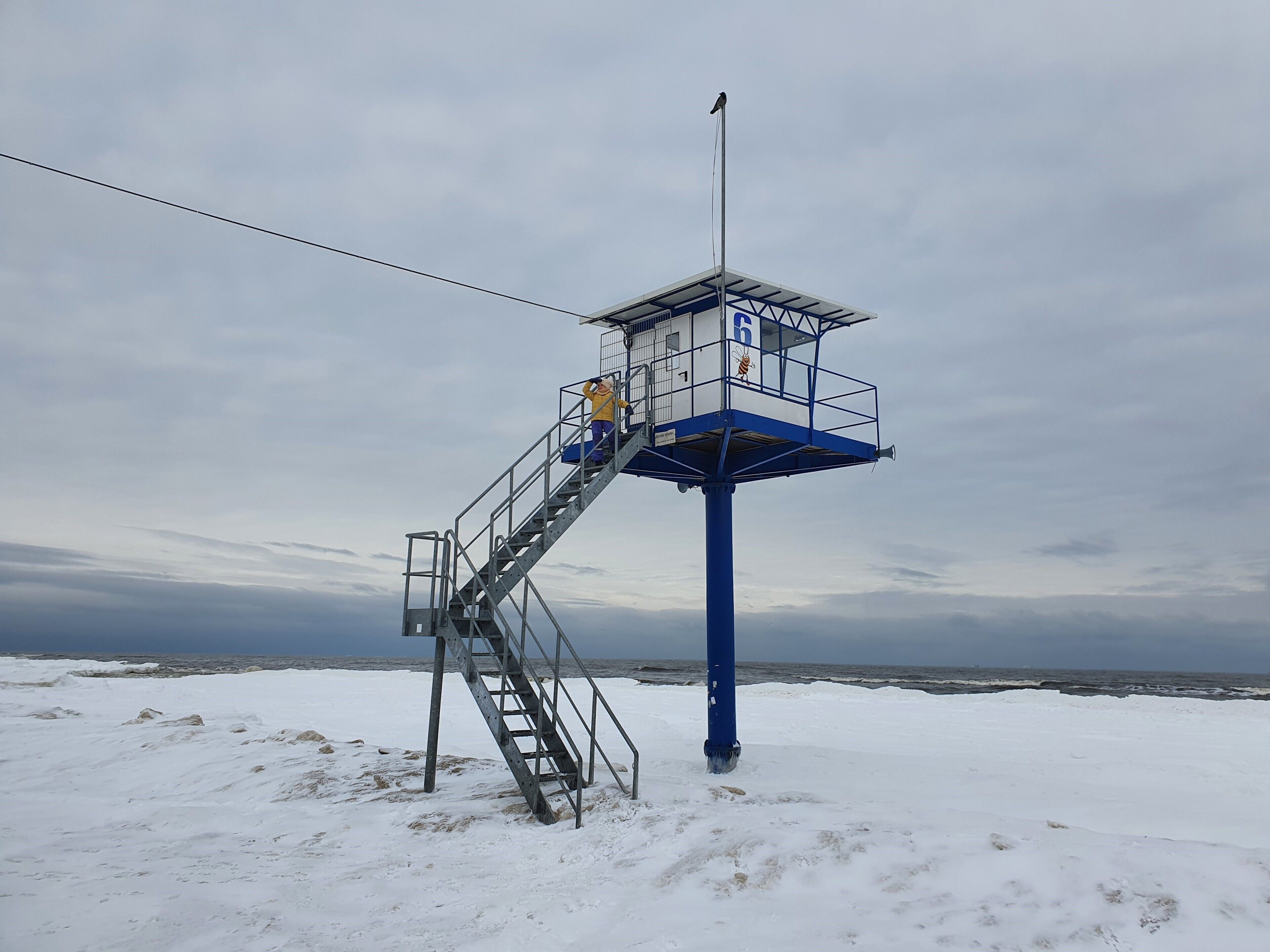 Ein Rettungsschwimmturm mit blauer Basis und weißem Oberteil, mit einer Fahnenmast und einer Treppe, die nach oben führt.