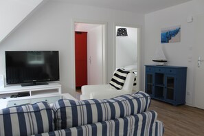 A living room with a TV on a white stand, a striped sofa, a white armchair, a blue cabinet, and a red door in the background.