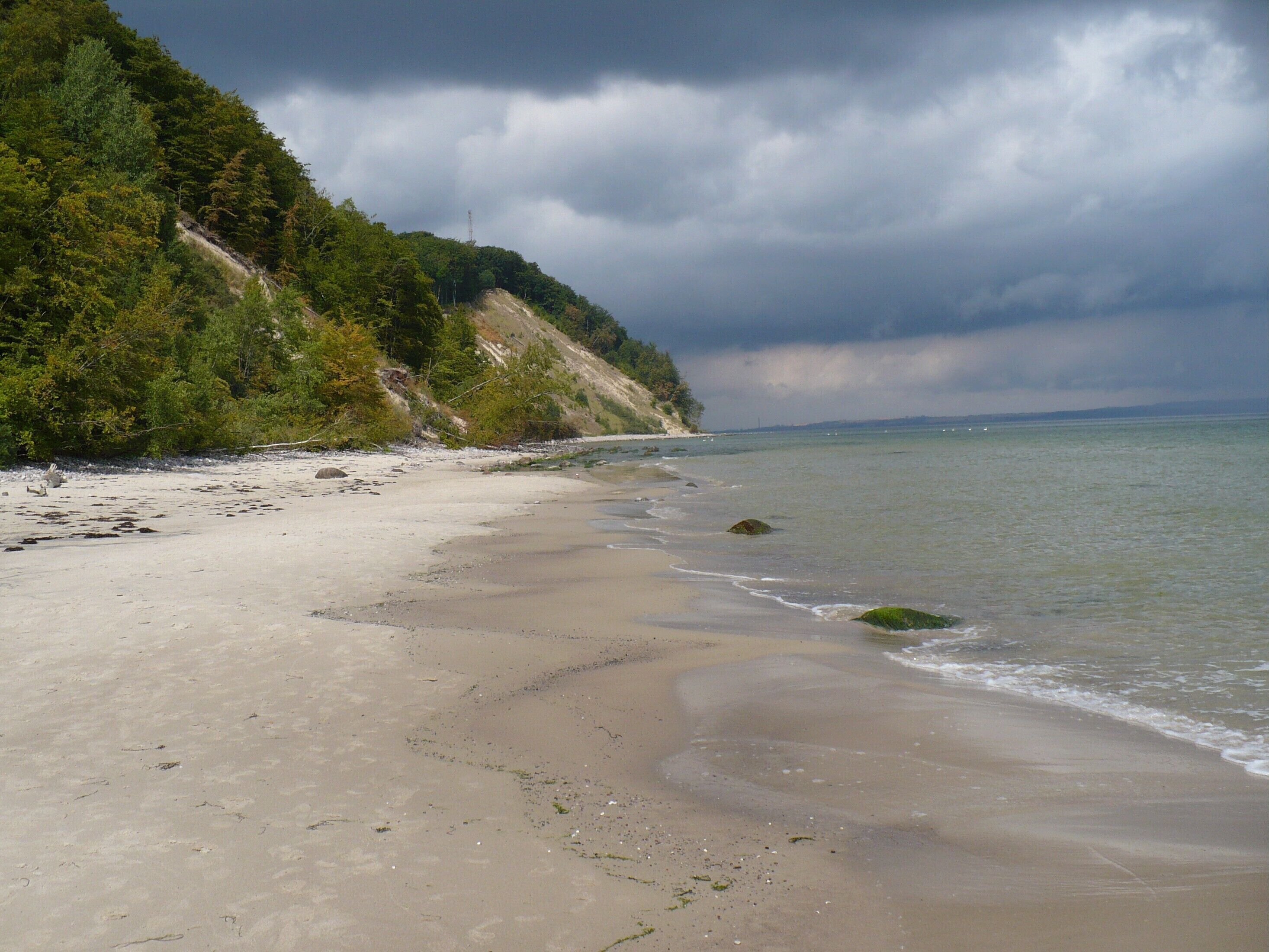 A beach with a sandy shore, a forested hillside, and a cloudy sky.