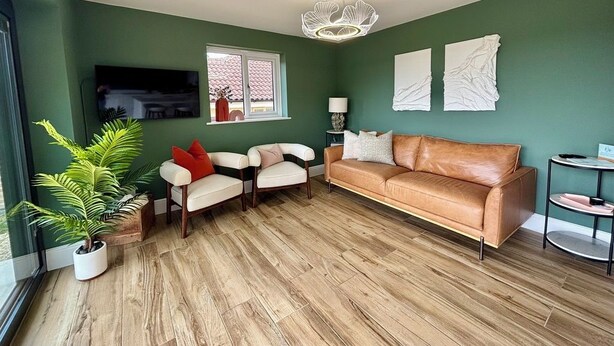 Sala de estar com parede verde, sofá de couro marrom, poltrona branca, piso de madeira e luminária no teto.