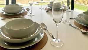 Mesa de jantar posta para quatro pessoas, com pratos, tigelas, talheres e copos, com vista para uma casa e vegetação.