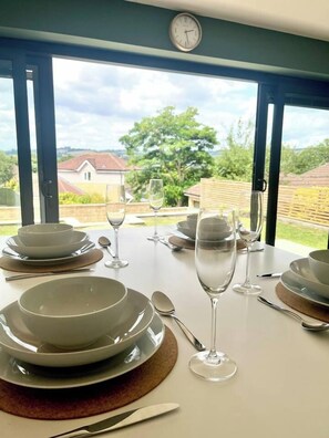 A dining table set for four with plates, bowls, cutlery, and glasses, with a view of a house and greenery outside. - 2bed Modern Home Garden Off Road Parking (Bath and North East Somerset)