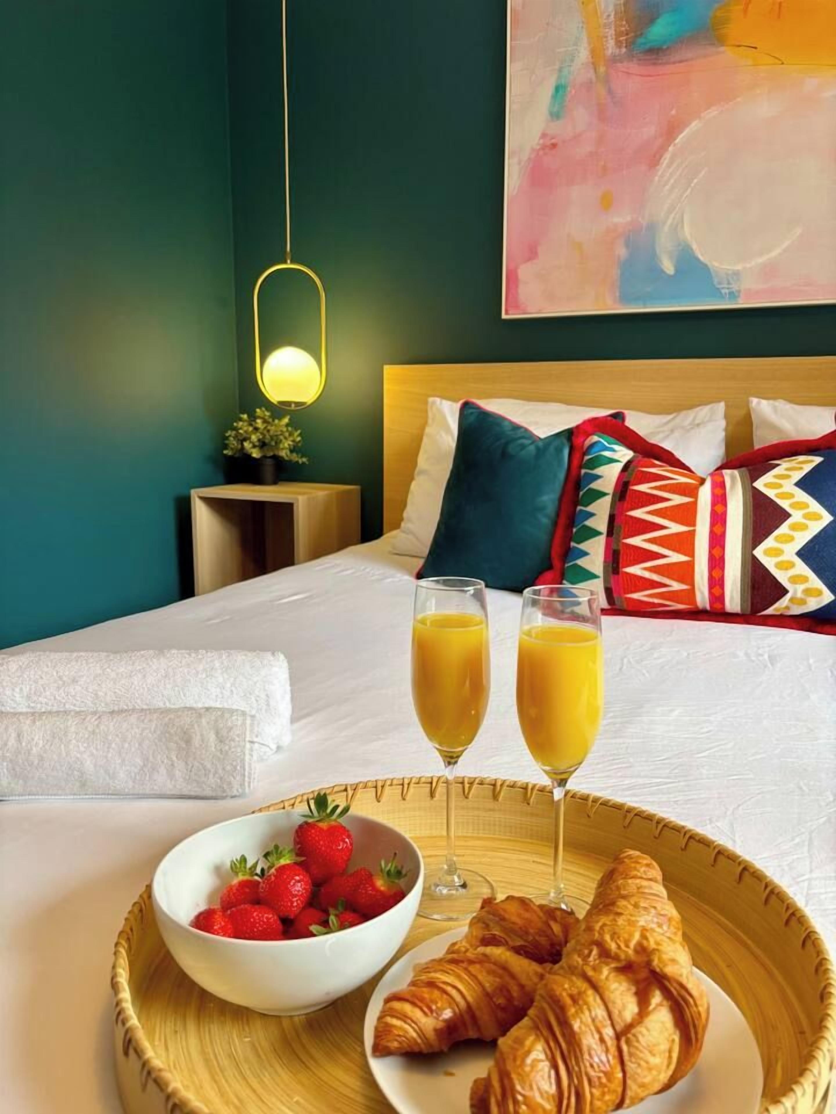Cama arrumada com edredom branco, bandeja com croissants e morangos, e dois copos de suco de laranja.