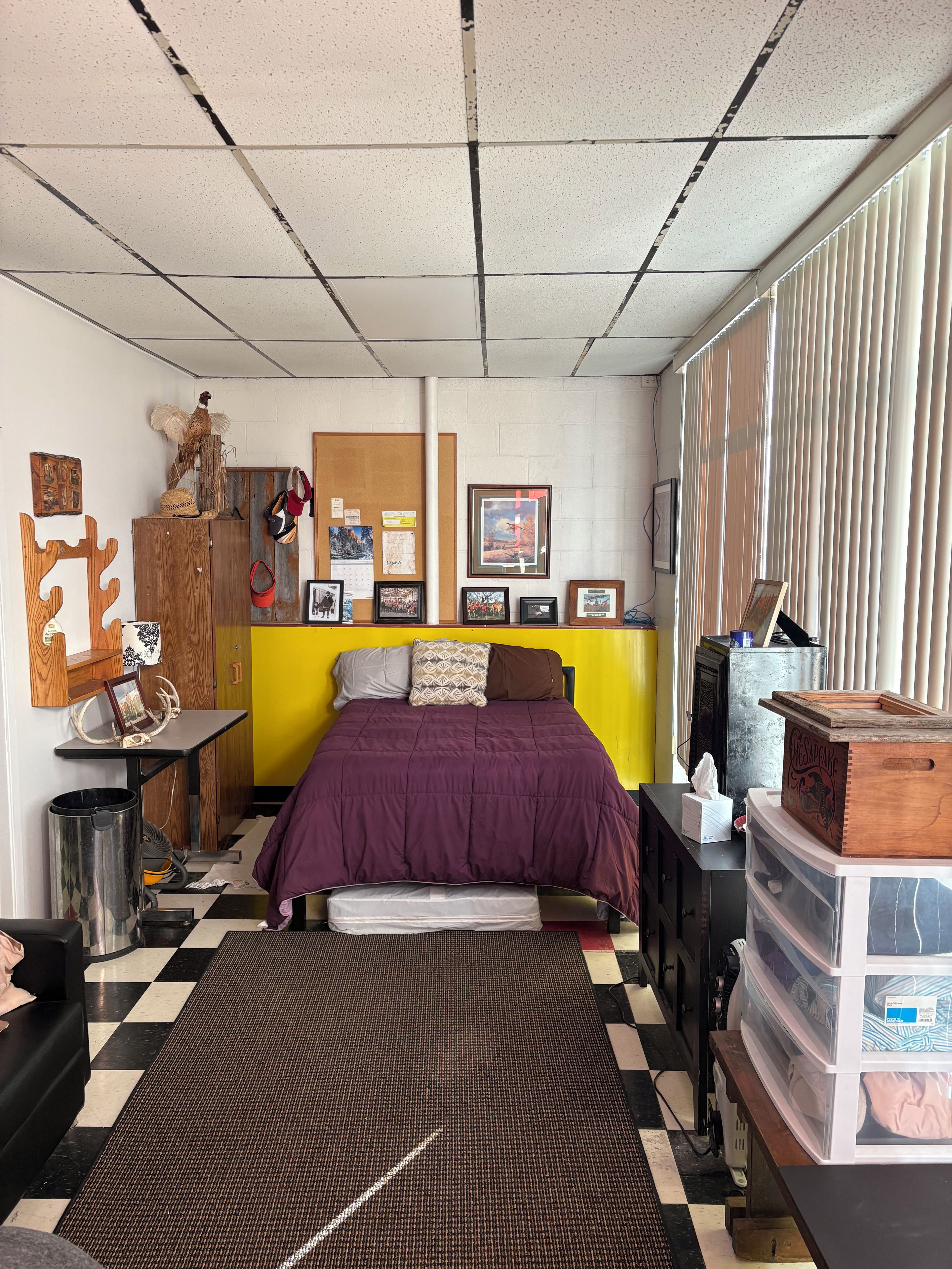 Un dormitorio con cama, un armario de madera, un cubo de basura, un radiador y una ventana con persianas.