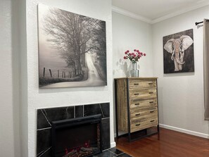 Een kamer met een open haard, een schilderij van een pad omzoomd door bomen, een kast met een bloevas, en een schilderij van een olifant.