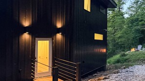 Eine dunkle Holzhütte mit einer Holztreppe, die zu einer Tür führt, umgeben von Kies und Bäumen.