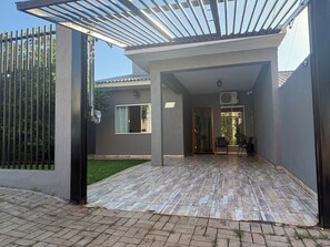 A covered outdoor area with a wooden floor, a metal gate, and a building with a window and an air conditioning unit. - Charming 2-bedroom house in serene Foz do Iguaçu with AC (Foz do Iguaçu)