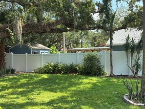 A residential backyard with a white fence, a house with a dark roof, and various plants. - Newly renovated beach house in quiet neighborhood, close to everything! (New Smyrna Beach)