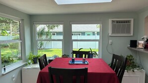 Un coin repas avec une table recouverte d’une nappe rouge, entourée de chaises. Deux fenêtres donnent sur un jardin.