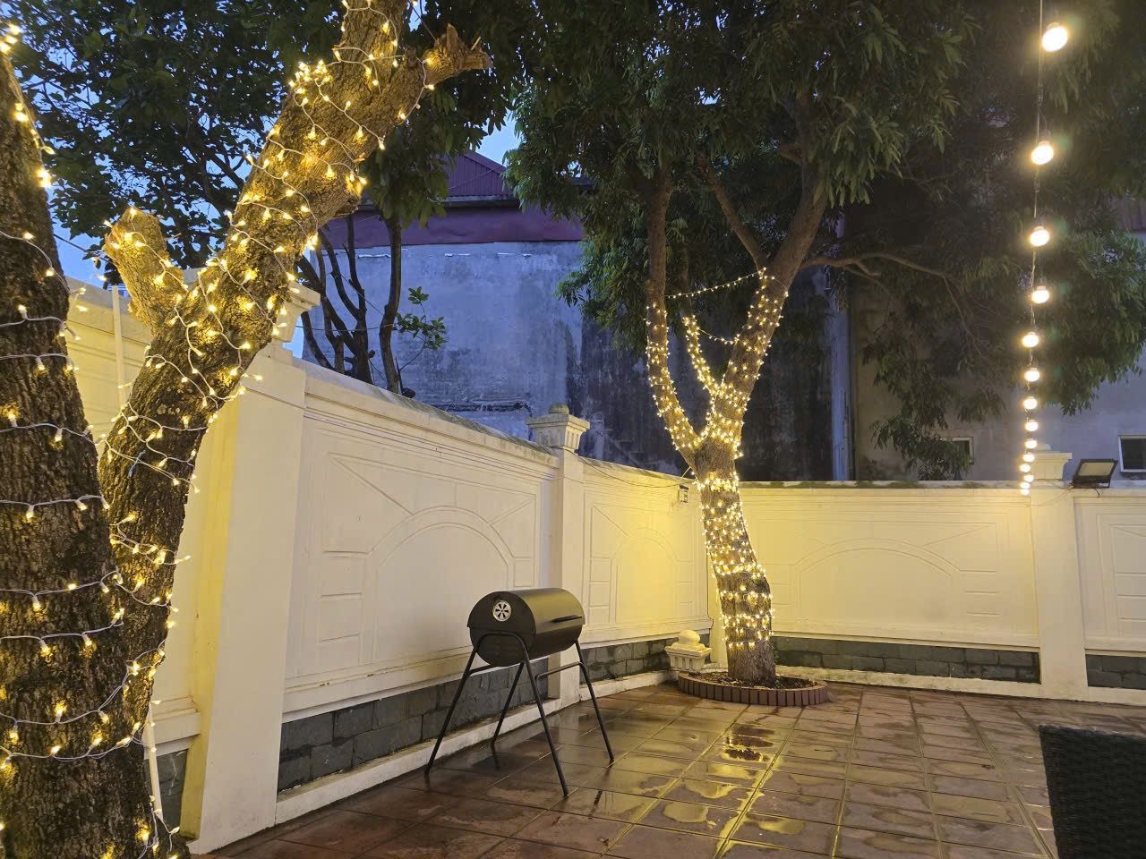 Eine Terrasse mit Grill, Bäumen, die mit Lichterketten umrankt sind, und einem weißen Zaun.