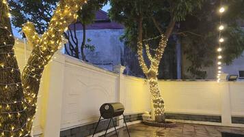 Eine Terrasse mit Grill, Bäumen, die mit Lichterketten umrankt sind, und einem weißen Zaun.