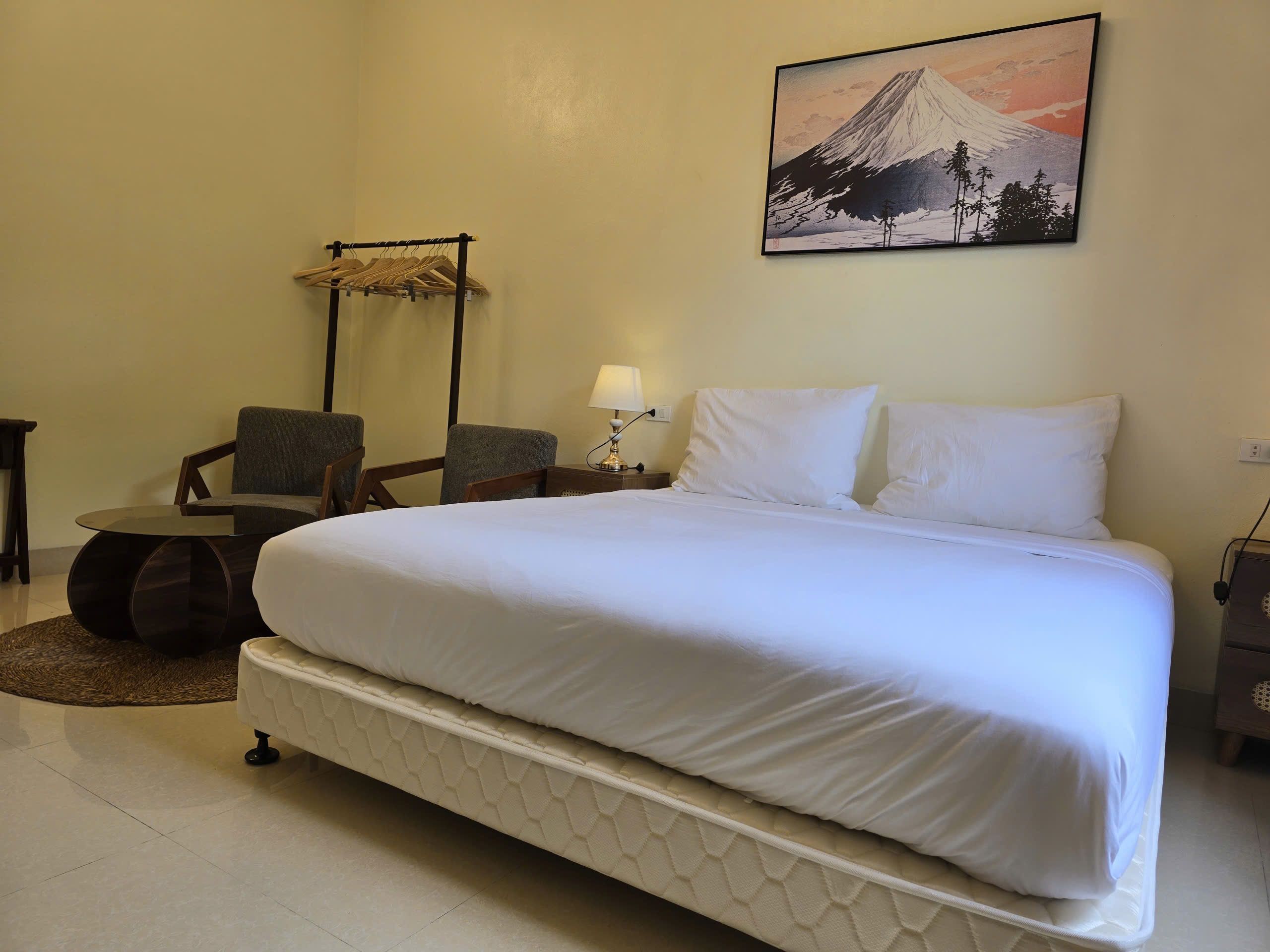 A hotel room with a large bed, a chair, a small table, and a wall-mounted lamp.