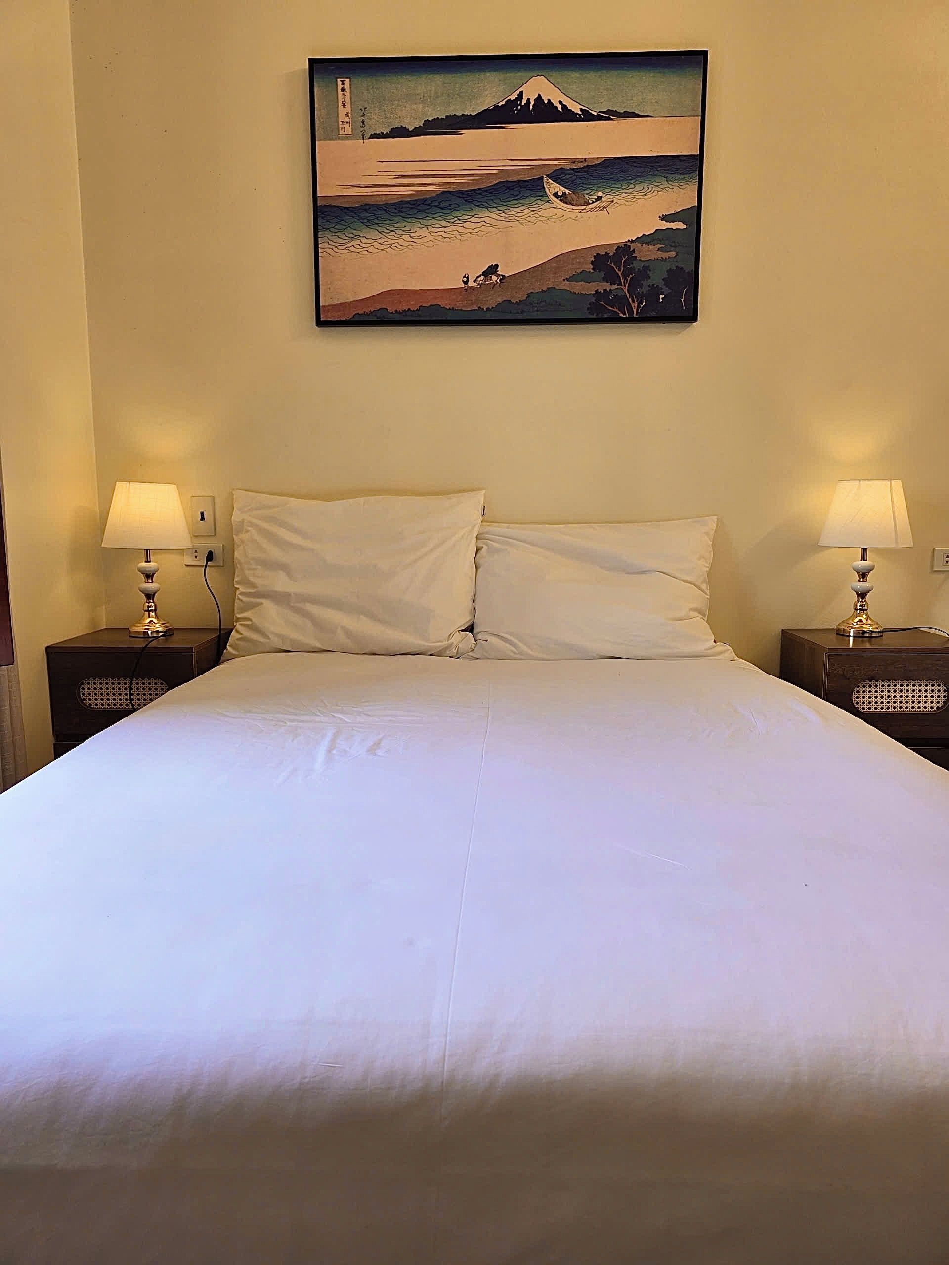 A bed with white linens, two bedside lamps, and a wall-mounted painting of a coastal scene.