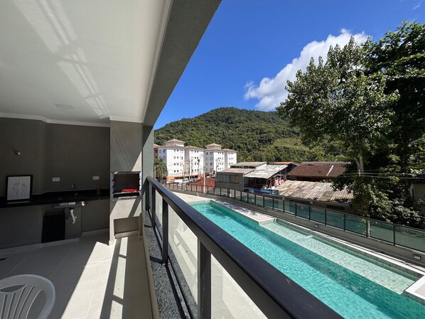 A balcony with a view of a swimming pool, a dining area, and a mountainous landscape in the background. - Brand new apartment. with complete leisure in the center (Ubatuba)