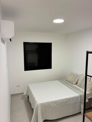 A bedroom with a bed, a window with closed blinds, a ceiling light, and a wall-mounted air conditioner. - Brand new apartment. with complete leisure in the center (Ubatuba)