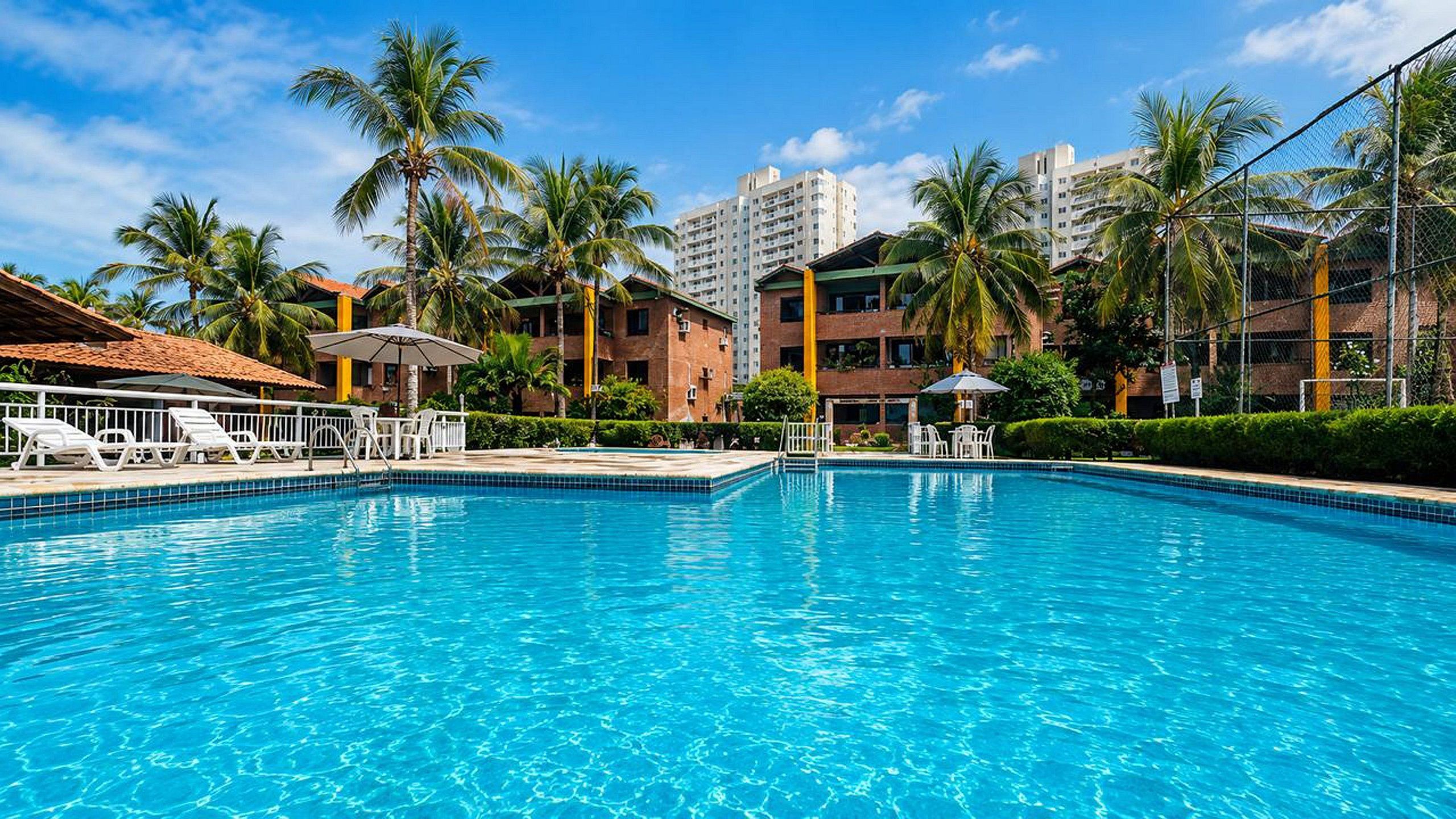 Una piscina con sillas de descanso y sombrillas, rodeada de palmeras y edificios.