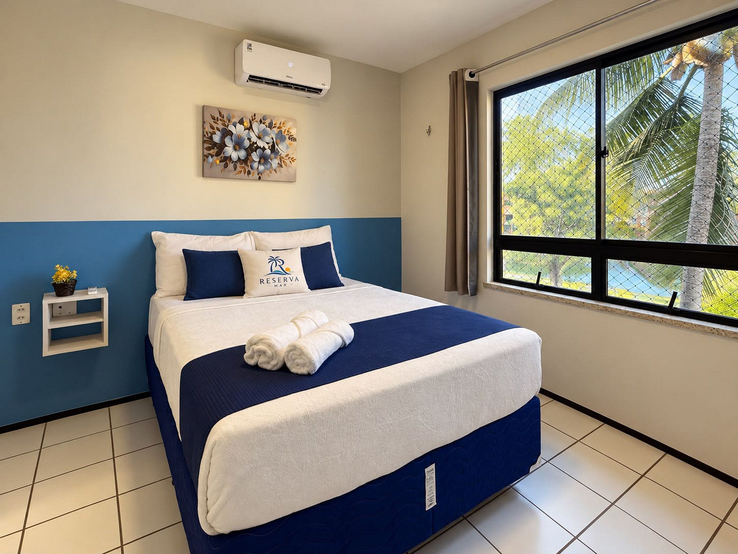 A hotel room with a bed, a wall-mounted air conditioner, a window with a view of palm trees, and a painting above the bed.