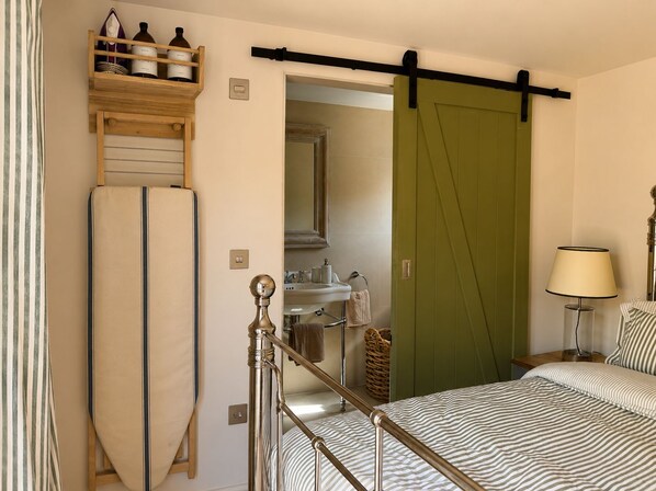 Un dormitorio con una cama, un estante con botellas, una puerta verde, una lámpara de noche y un baño visible a través de una puerta abierta.