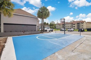Un terrain de basketball extérieur, doté d’une surface bleue, entouré d’une clôture et situé à côté d’un bâtiment à la façade grise.