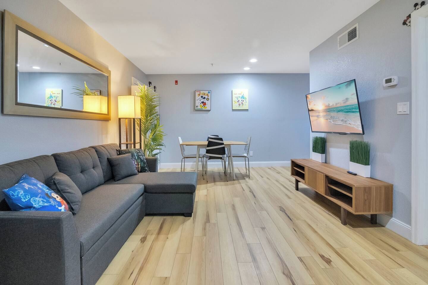A modern living room with a sectional sofa, a dining table with chairs, a TV on a wooden stand, and a large mirror on the wall.