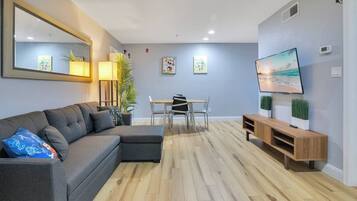 A modern living room with a sectional sofa, a dining table with chairs, a TV on a wooden stand, and a large mirror on the wall.