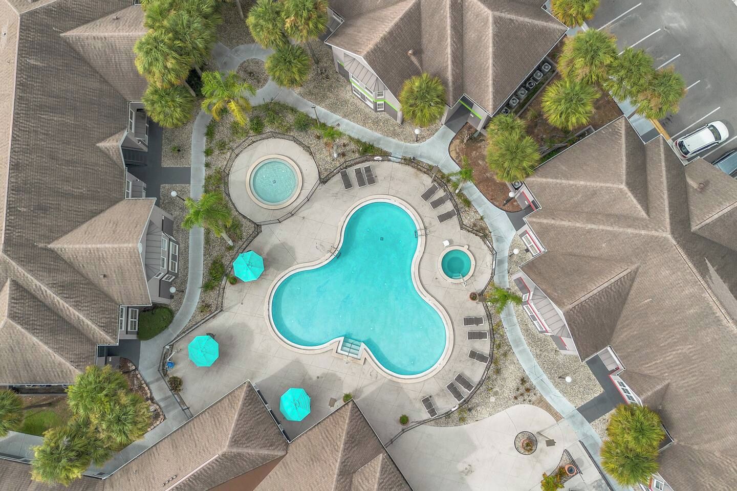 A residential area with a large swimming pool surrounded by lounge chairs and umbrellas, with multiple houses in the background.