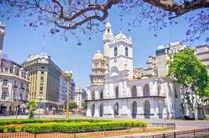 Un edificio histórico con una torre de reloj, rodeado de arquitectura moderna y un jardín bien cuidado.
