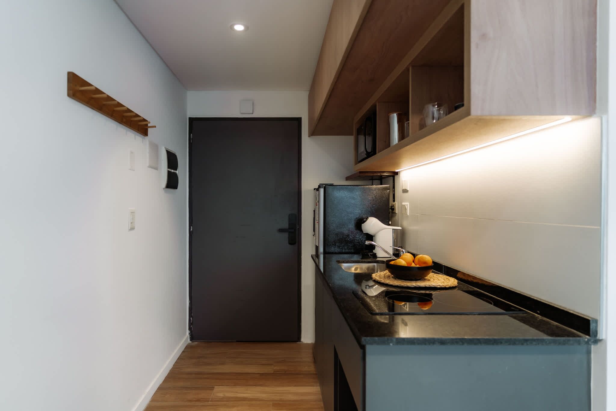 A narrow kitchen with a dark door, wooden shelves, and a countertop with a bowl of fruit.
