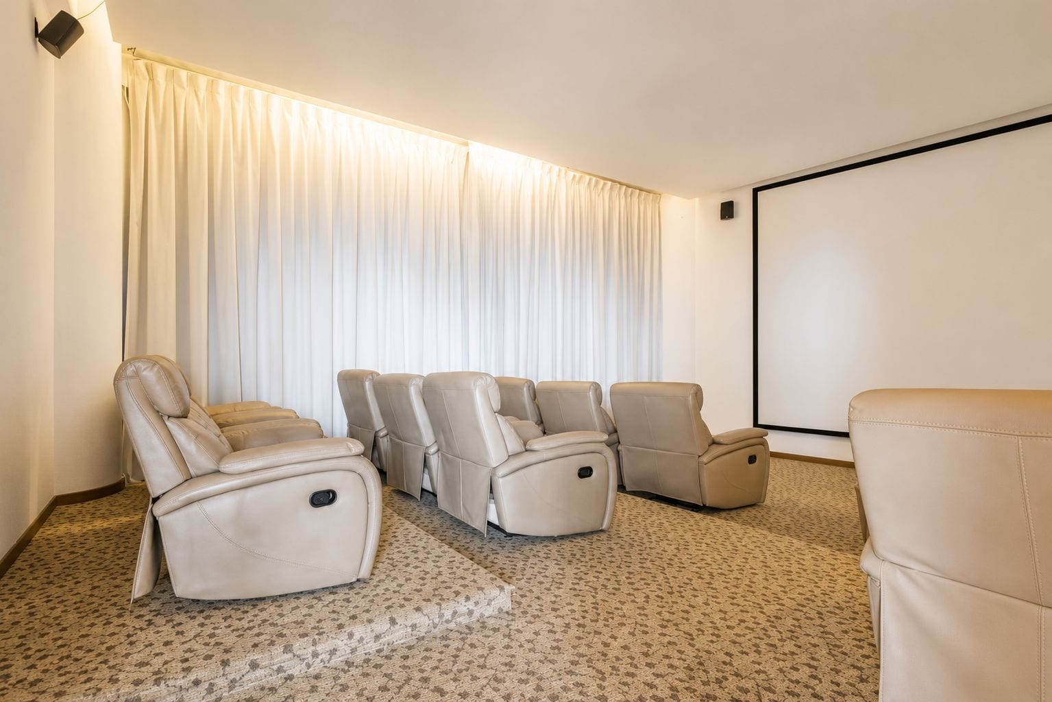 Un salón de cine en casa con cuatro butacas reclinables, piso alfombrado, una cortina blanca grande y una pantalla de proyección vacía.