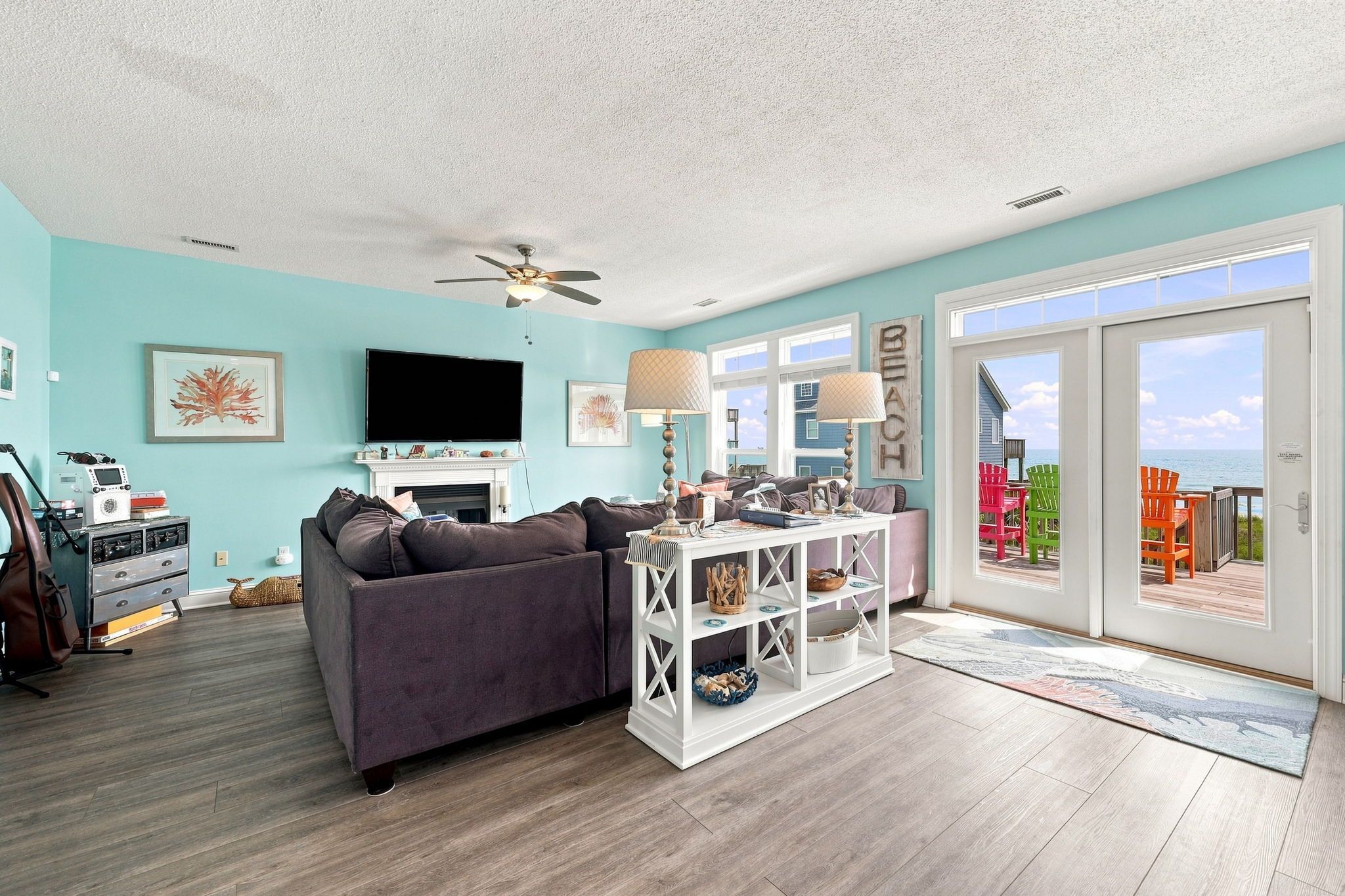 A living room with a sofa, a TV, a ceiling fan, and a view of the beach through a glass door.