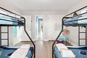 A room with two bunk beds, a small table with a chair, and a view of a hallway leading to a door. - Almost Heaven | Beachside Living, Ocean Breezes (North Topsail Beach)
