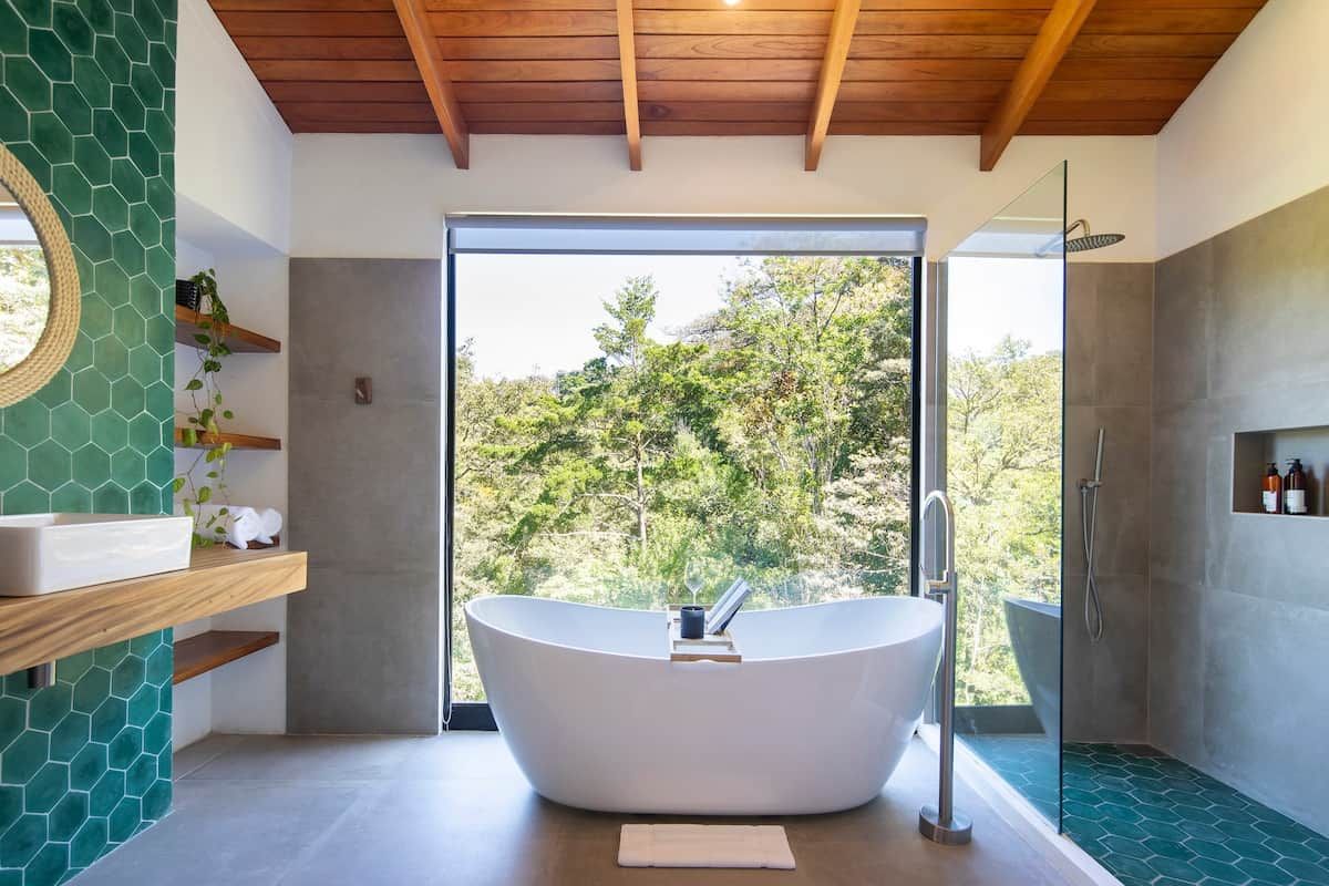 Un baño moderno con una bañera independiente, una ducha con cerramiento de vidrio y vistas a árboles a través de un ventanal grande.