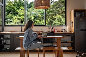 A person sitting at a wooden table with a laptop, in a room with a large window overlooking greenery.