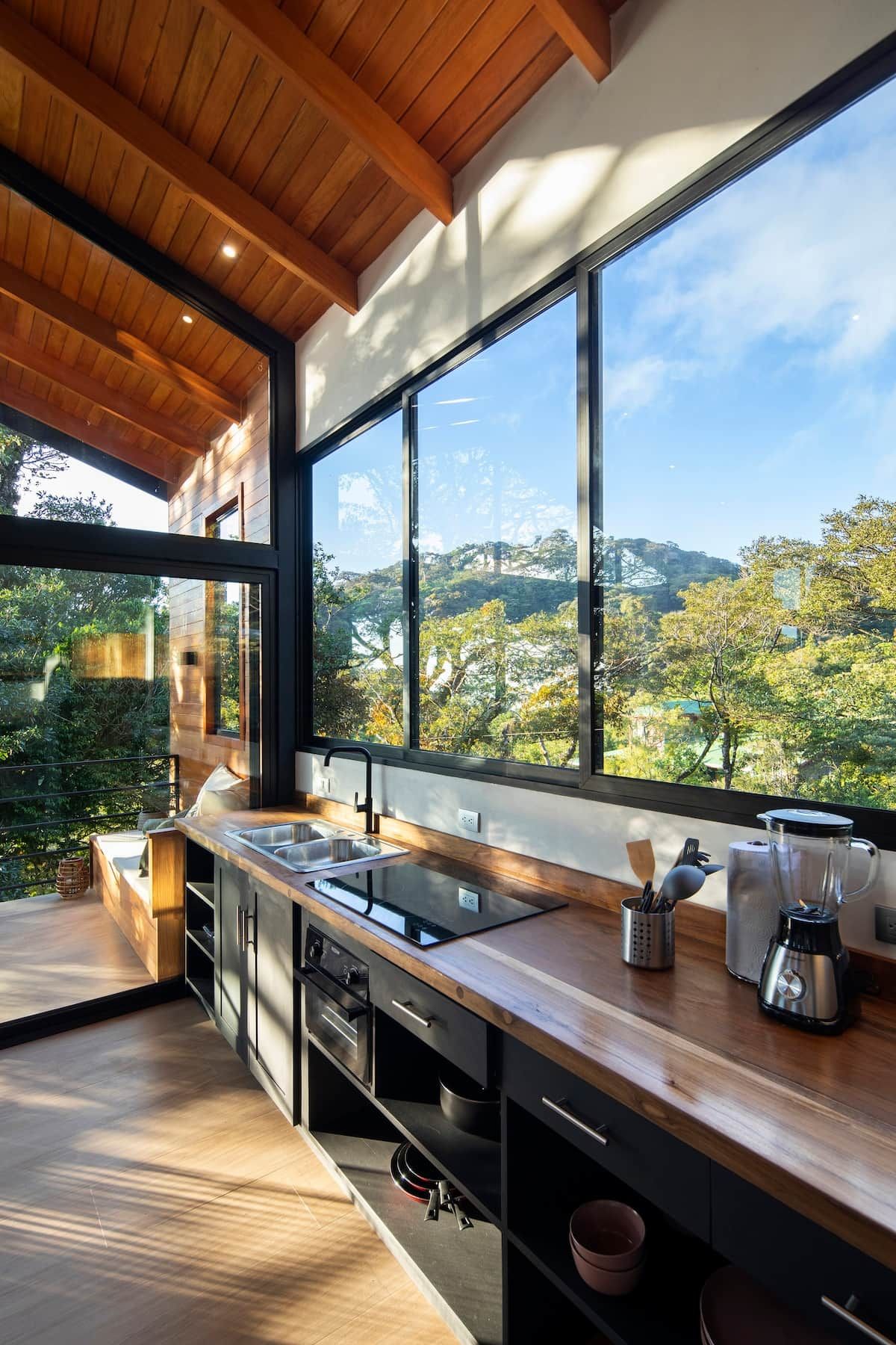 Eine moderne Küche mit großem Fenster, Holzboden und Blick auf eine bewaldete Landschaft.
