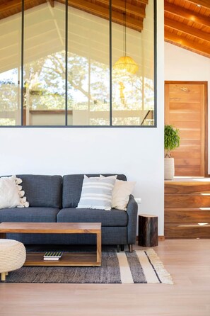A modern living room with a grey sofa, a wooden coffee table, and a large window with a view of trees. - Casa Jaguar (2) (Monteverde)